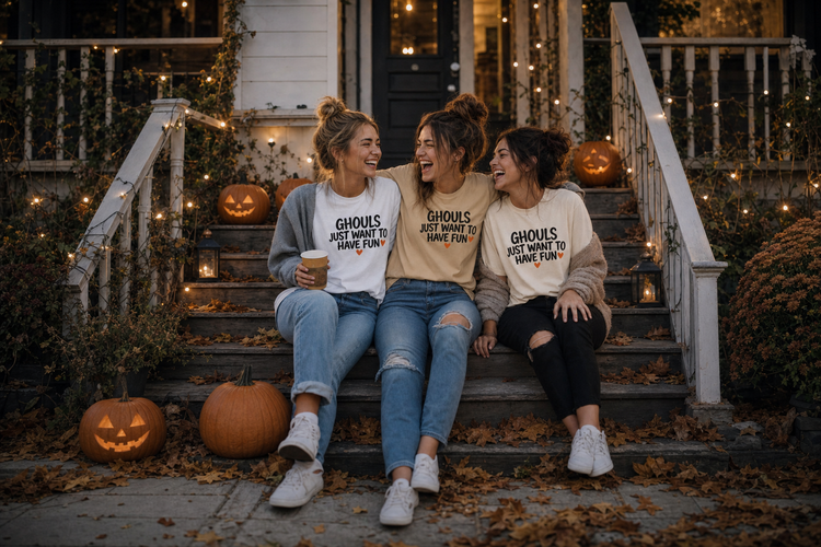 Three friends laughing on autumn porch steps wearing the Ghouls Just Want To Have Fun tee, OMG Halloween range