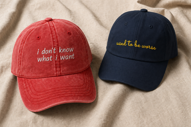 Two embroidered caps on oatmeal linen: a washed red vintage cap reading “i don't know what i want” in white, and a navy classic cap reading “used to be worse” in gold